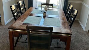 Une salle à manger avec une table en bois, quatre chaises, un vase de fleurs et une vue dégagée sur l’extérieur grâce à de grandes fenêtres ornées de rideaux.