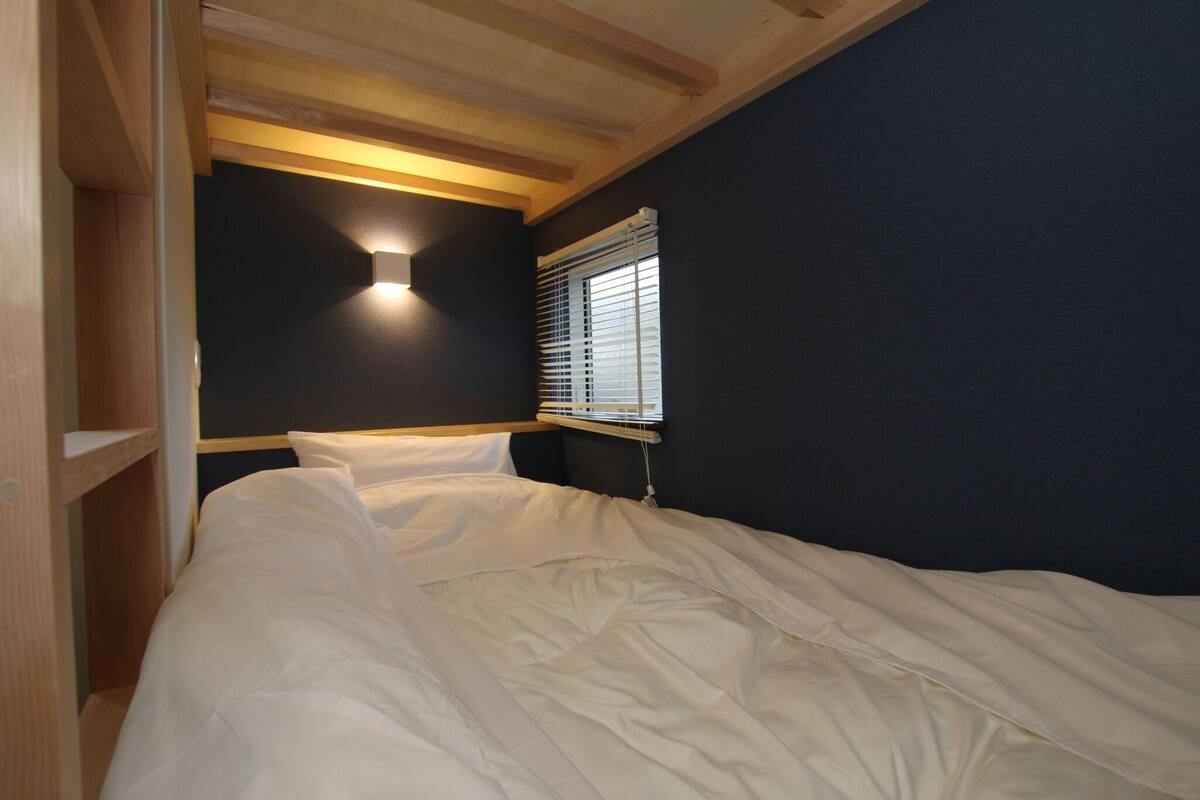 A bedroom with a bed, a nightstand, a wall-mounted light, and a window with blinds.