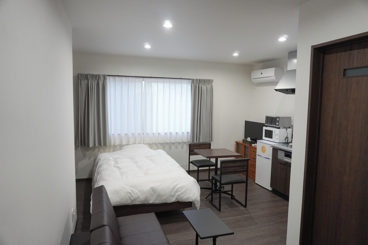 A hotel room with a bed, a desk, a chair, a small table, a microwave, and a kitchenette.