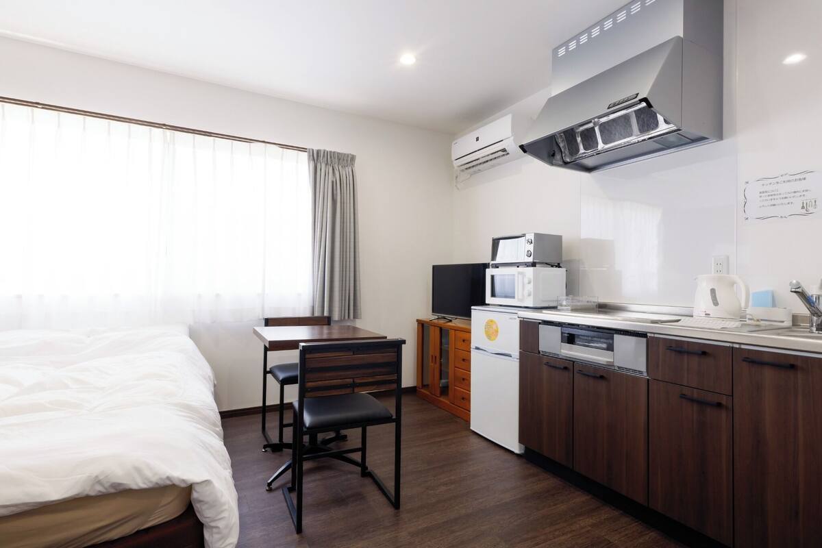 A compact hotel room with a bed, a desk, a kitchenette, and a window with curtains.