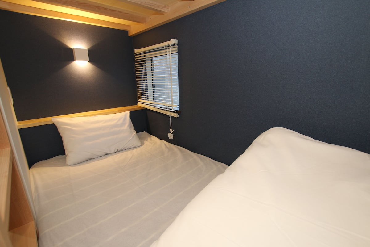 A single bed with white bedding, a wall-mounted light, and a window with blinds.