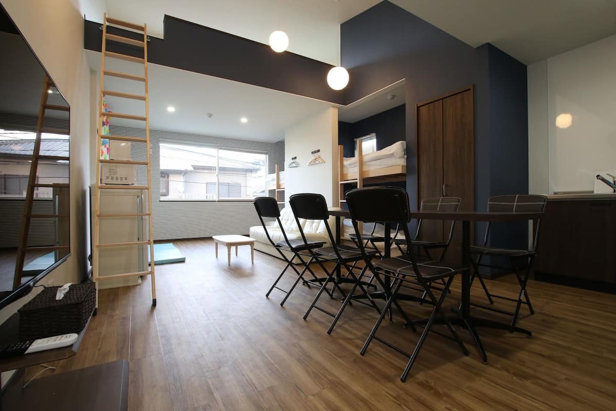 A room with a wooden floor, a dining table with chairs, a sofa, a ladder, and a window with blinds.