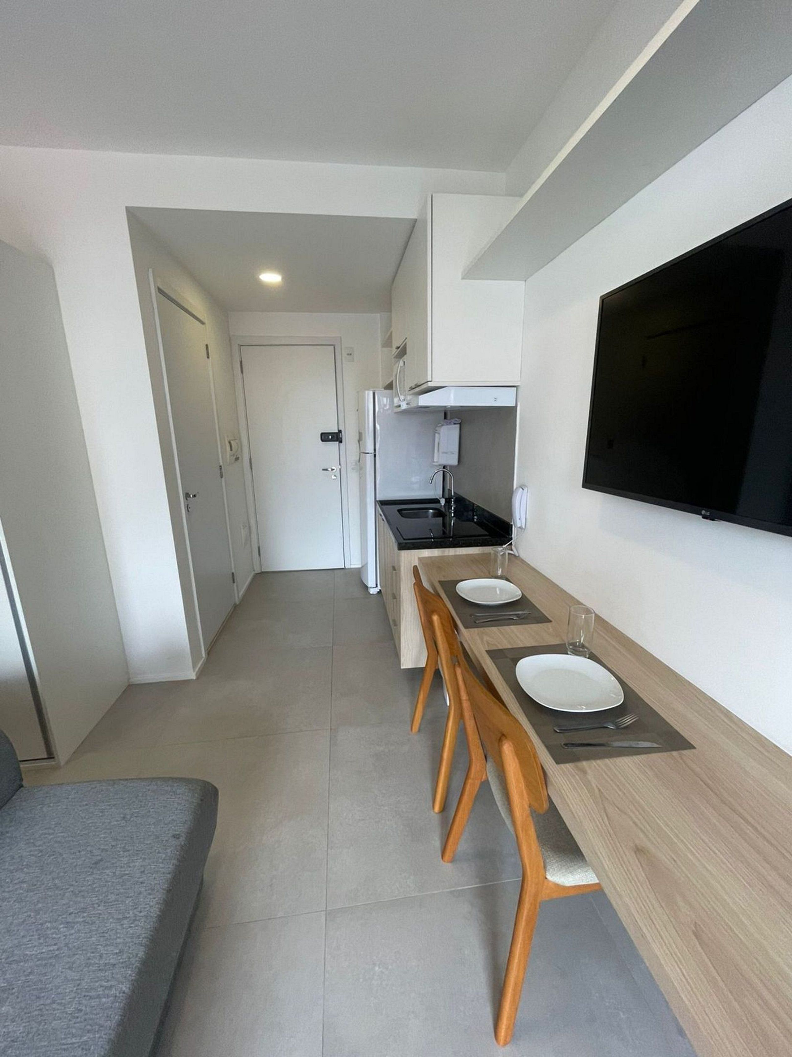 A modern kitchen with a dining area, a TV mounted on the wall, and a door leading to another room.