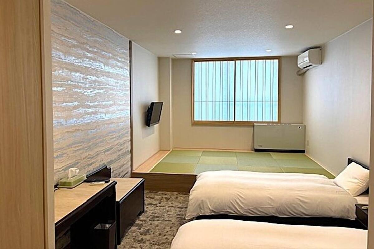A hotel room with two beds, a desk, a TV, and a large window with blinds.