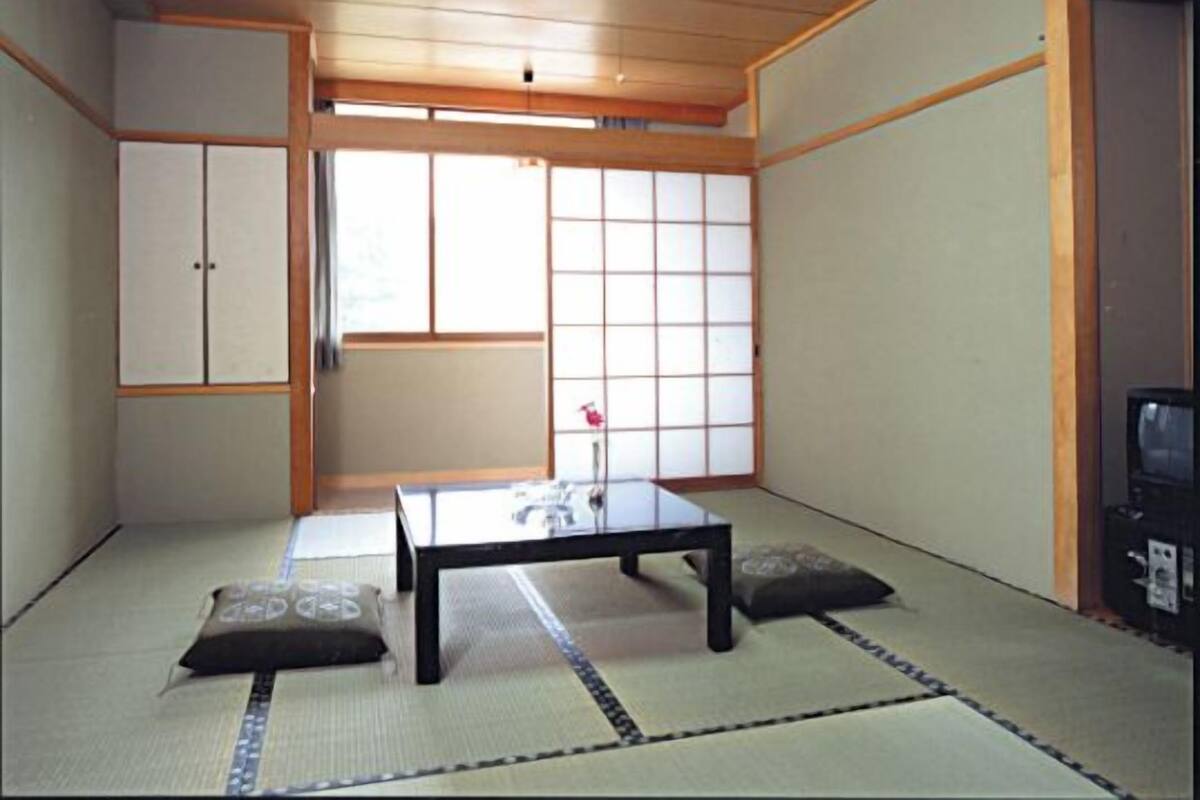 A traditional Japanese room with tatami flooring, a low table, cushions, sliding doors, and a television.
