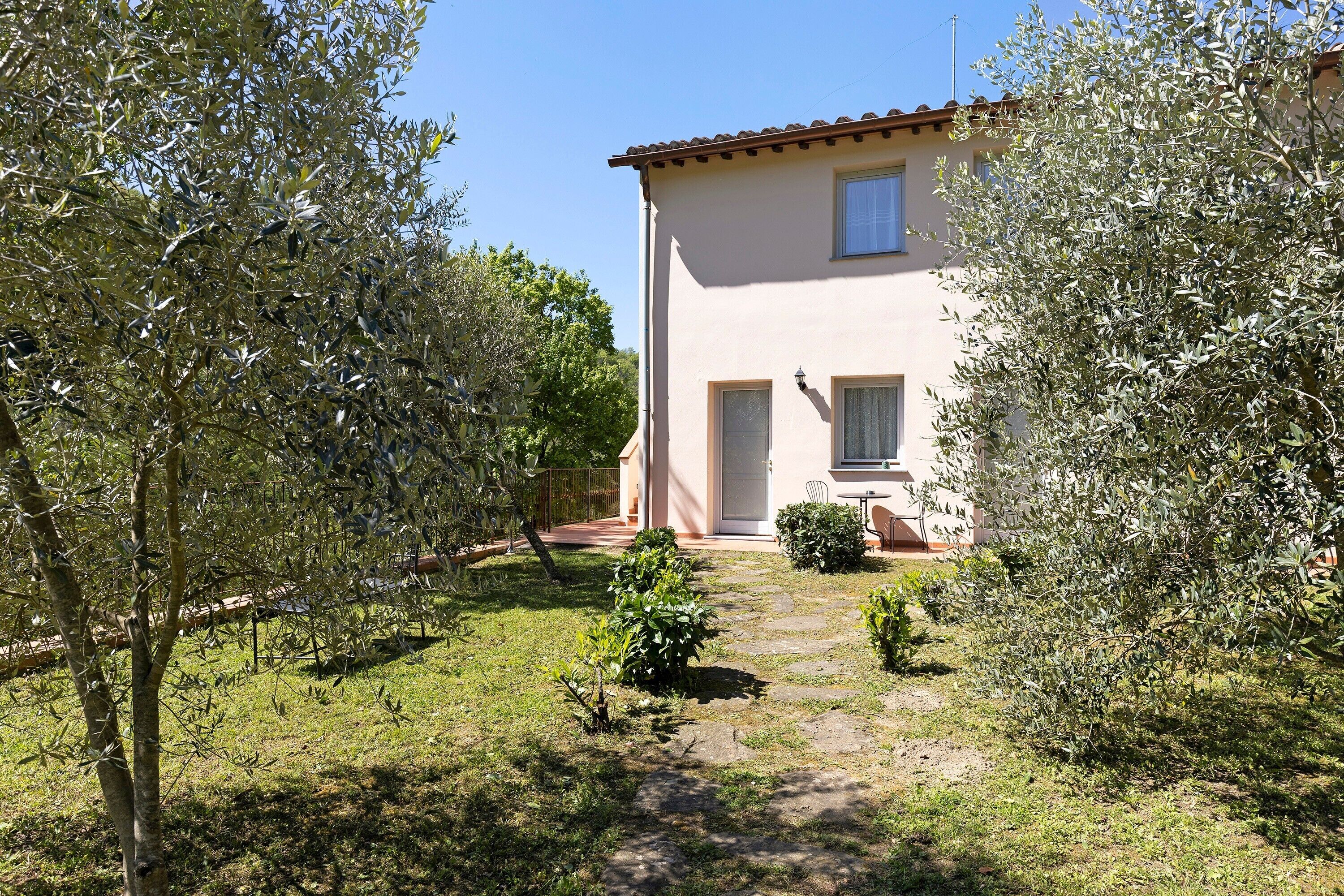 A two-story house with a stone pathway leading to the entrance, surrounded by olive trees and a small garden.