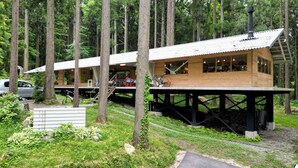 A cabin with a wooden exterior, a metal roof, and a deck. There is a car parked outside and a bicycle leaning against the cabin. - Morinonakano Kissatooyado Morikurasu (Maniwa)