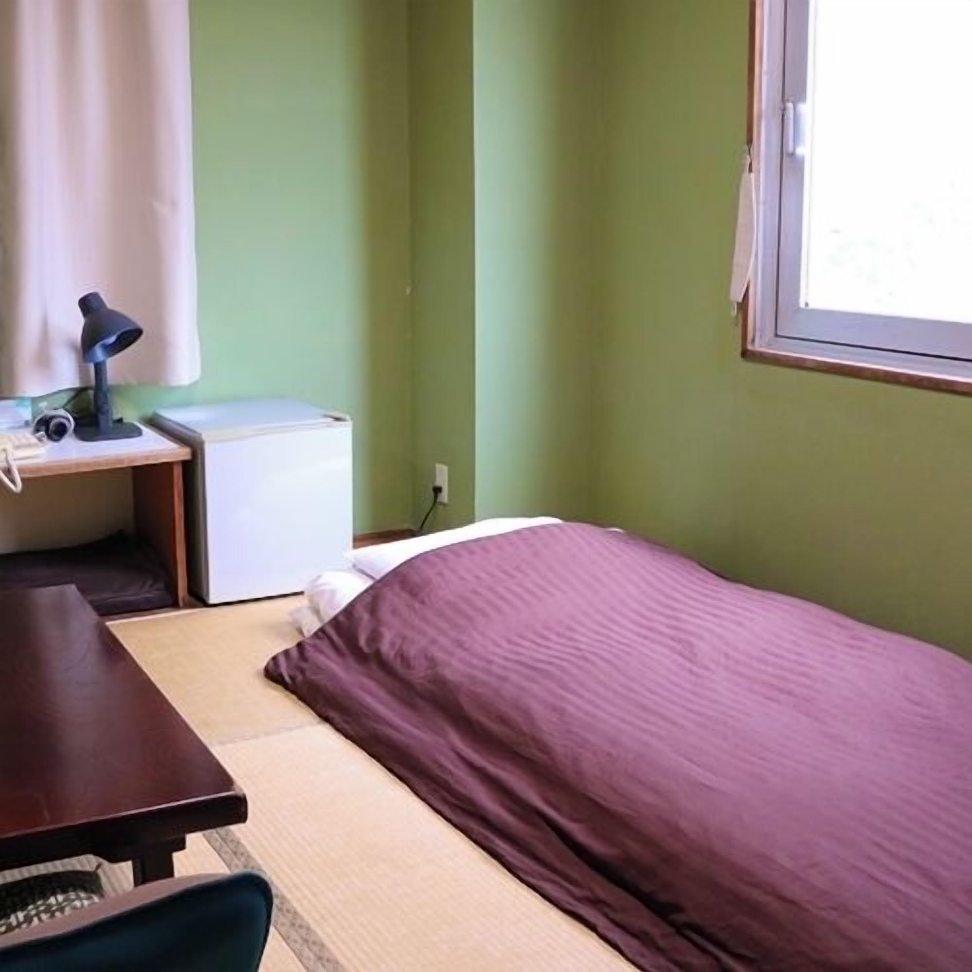 Quarto com cama, escrivaninha com luminária, eletrodoméstico pequeno e janela.