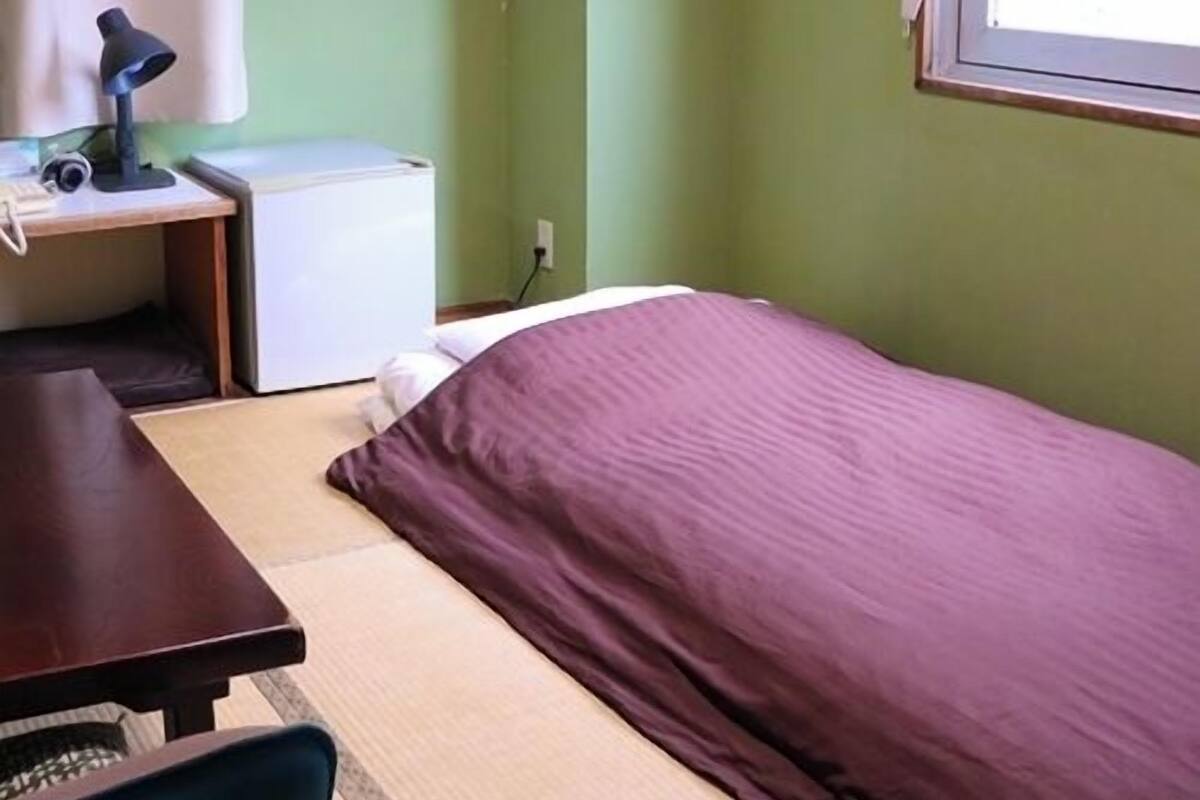 A room with a bed, a desk with a lamp, a small white appliance, and a window.