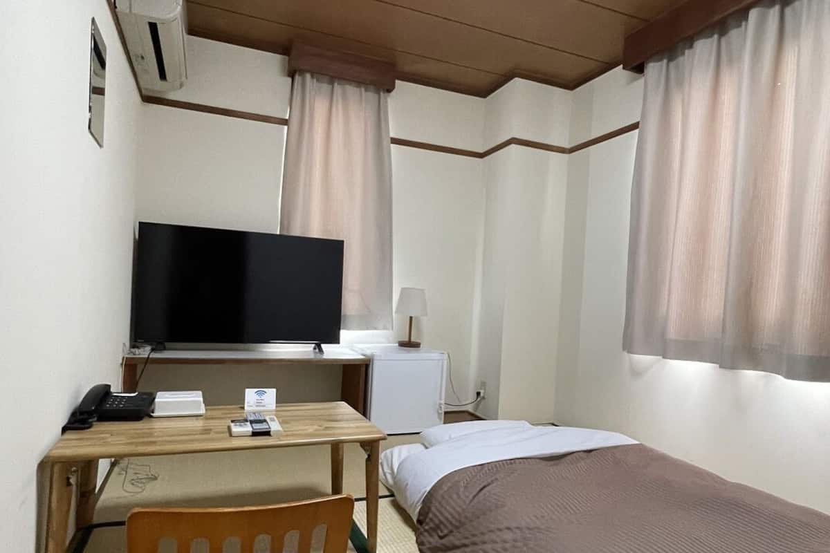 A compact hotel room with a single bed, a desk with a computer and phone, a TV, and a small bedside table.