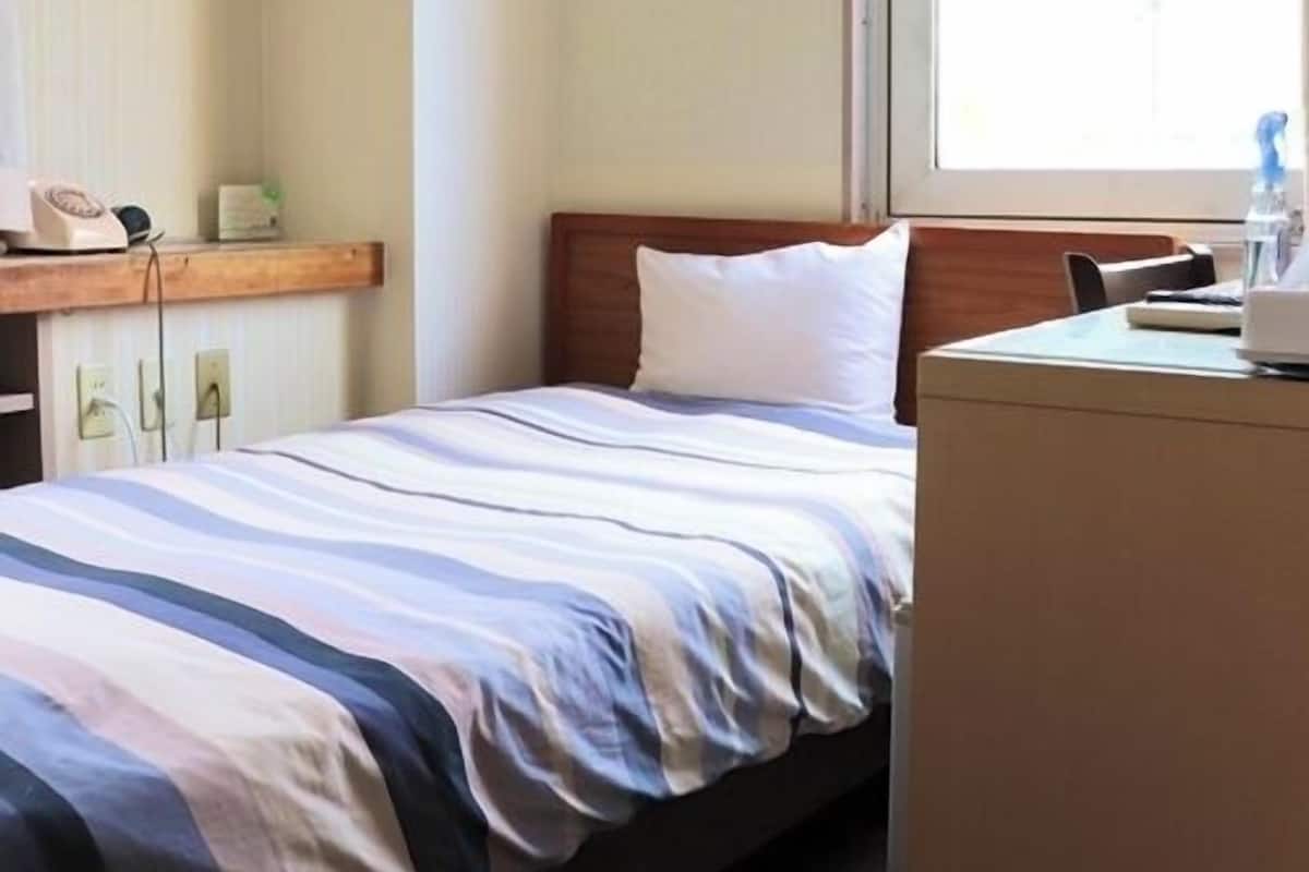 A single bed with a striped bedspread, a wooden headboard, a bedside table with a lamp, and a window with blinds.