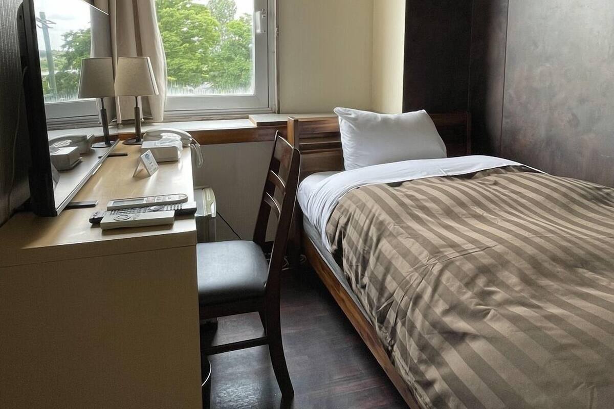 Quarto de hotel com cama, escrivaninha, cadeira, abajur e janela com cortinas.