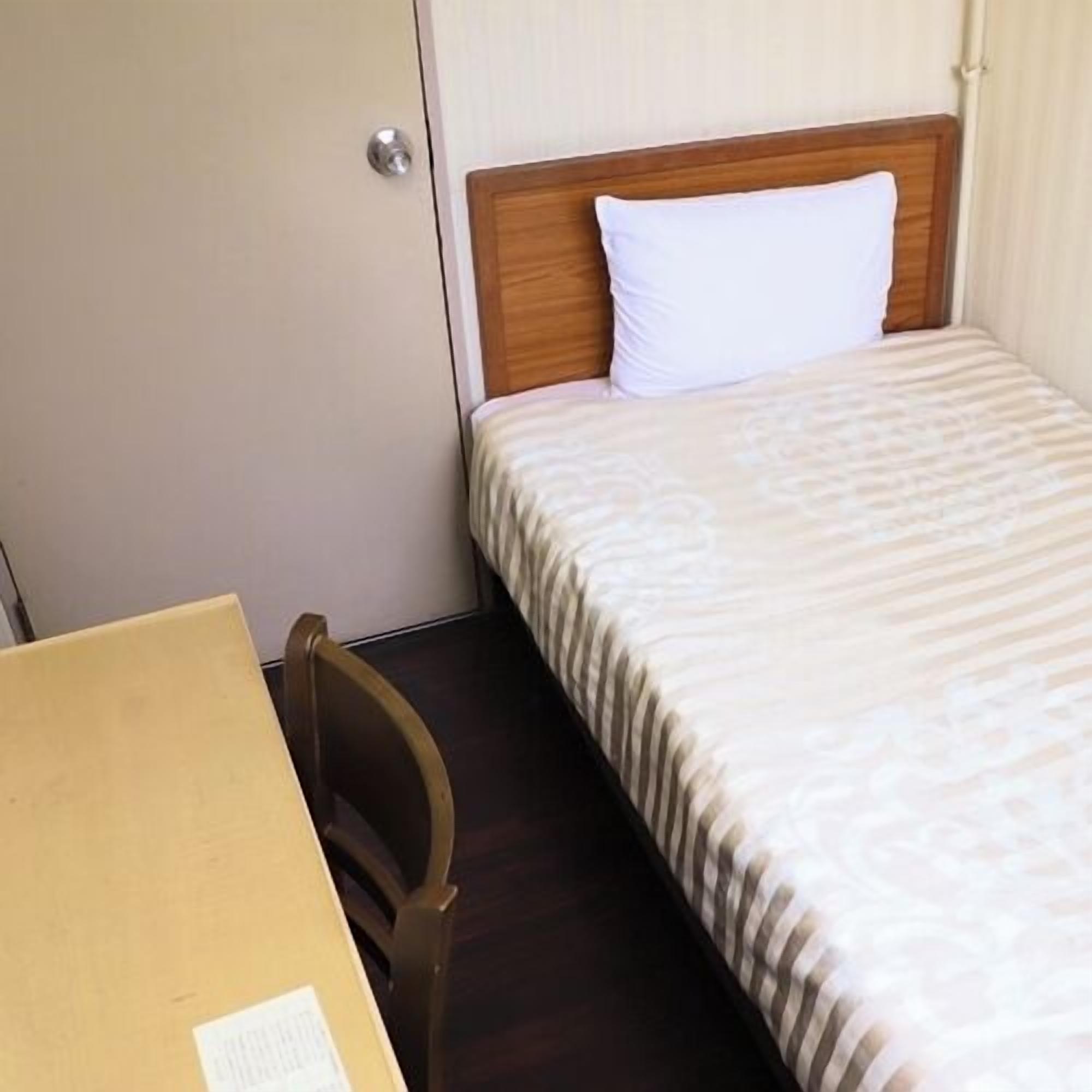 Uma cama de solteiro com cabeceira de madeira, travesseiro branco e colcha estampada. Há uma cadeira de madeira e uma mesinha com um papel em cima.