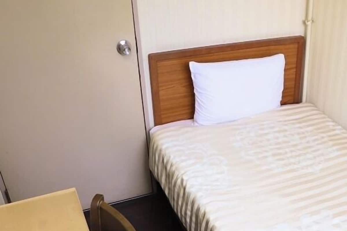 A single bed with a wooden headboard, a white pillow, and a patterned bedspread. There is a wooden chair and a small table with a paper on it.
