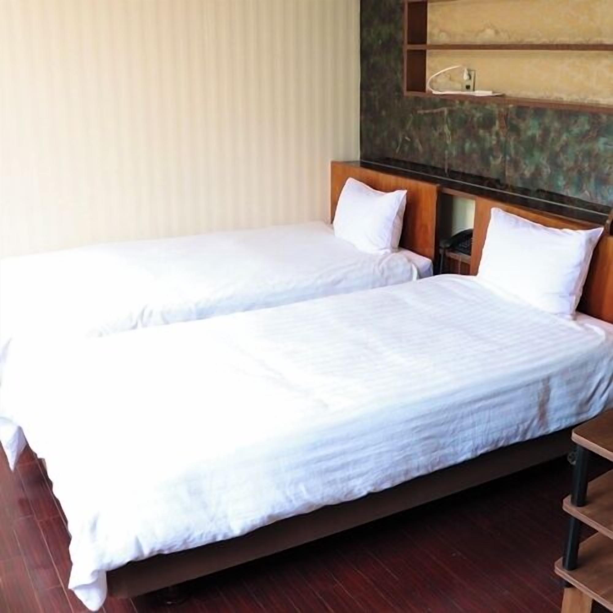 Quarto de hotel com duas camas de solteiro, cabeceiras de madeira, uma mesa de cabeceira e uma prateleira fixada na parede.