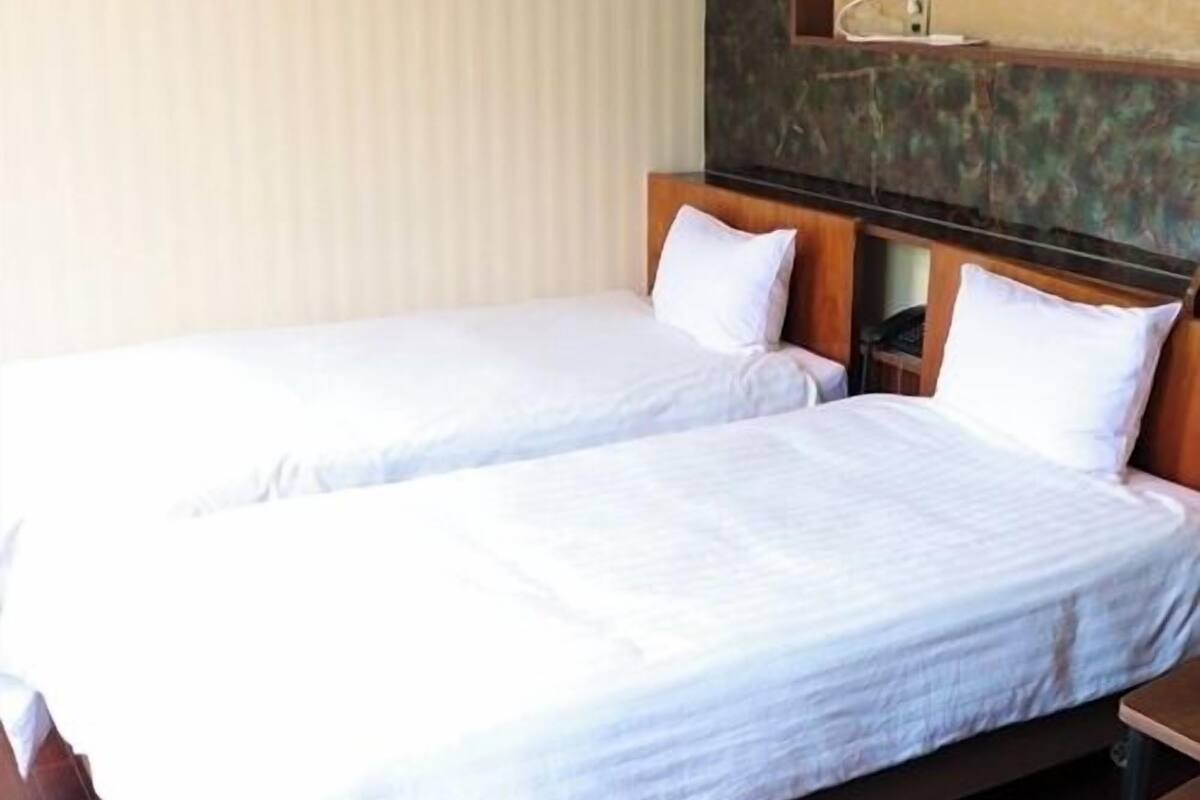 Quarto de hotel com duas camas de solteiro, cabeceiras de madeira, uma mesa de cabeceira e uma prateleira fixada na parede.