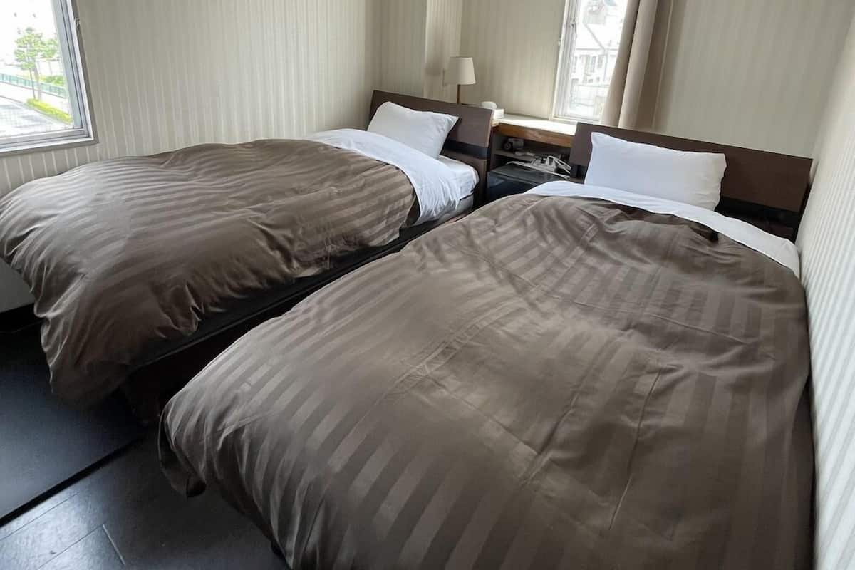 A hotel room with two beds, a nightstand, a lamp, and a window with curtains.