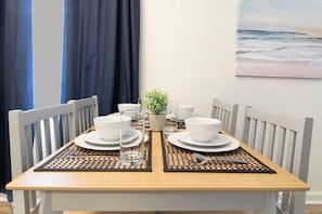 A dining table set with white plates, bowls, and glasses, surrounded by white chairs, with a view of the ocean through a window. - Centrally Located- The Delightful Doubles Unit E (Columbus)