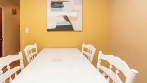 Een eetkamer met een witte tafel en stoelen, een gele muur en een abstract kunstwerk.