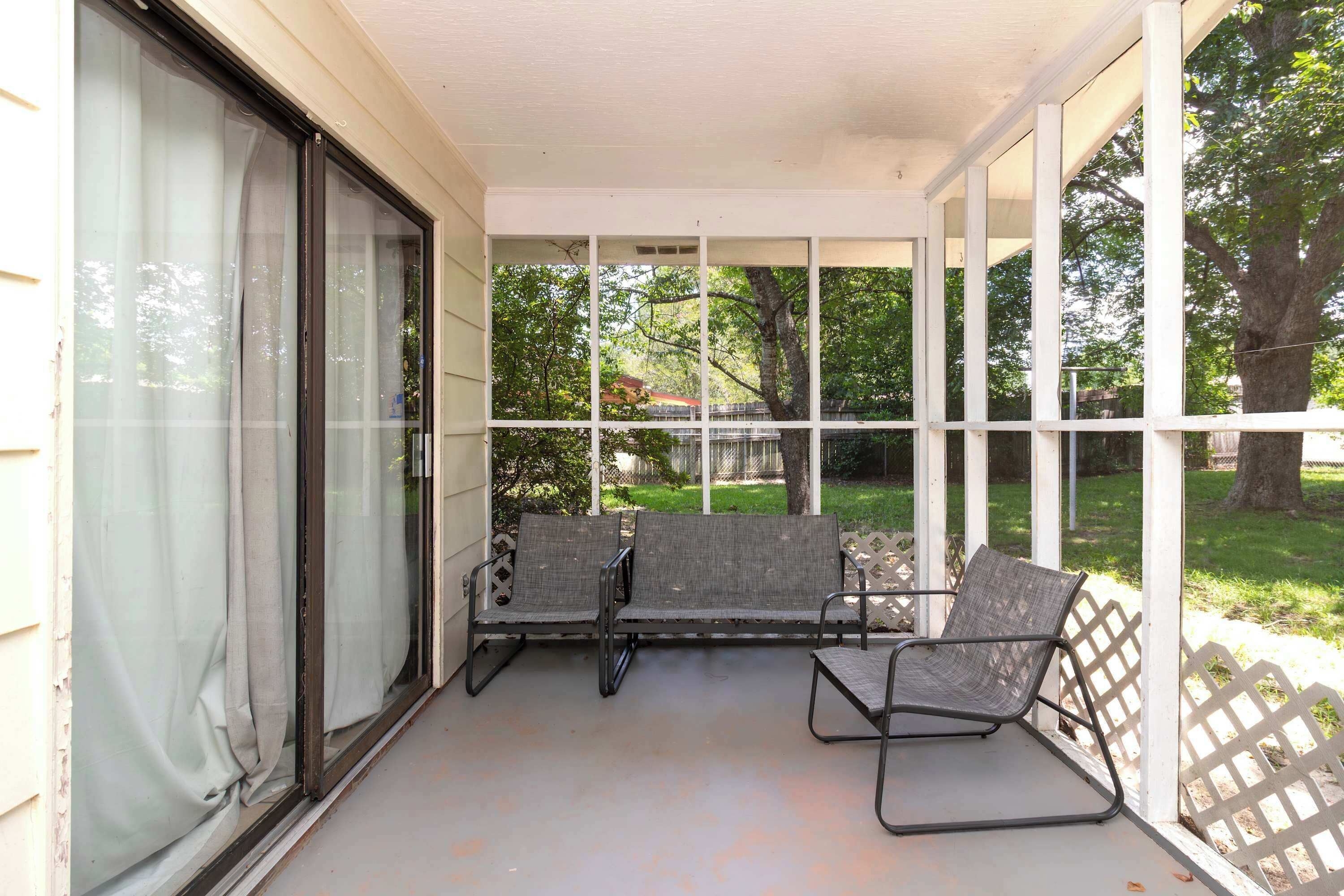 Eine überdachte Veranda mit zwei Stühlen und einer Bank, große Fenster und Blick auf einen Garten mit Bäumen.