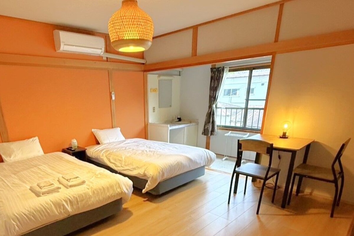 Une chambre avec deux lits, un bureau et une chaise. Il y a une fenêtre avec des rideaux, un luminaire suspendu et une petite table avec une lampe.