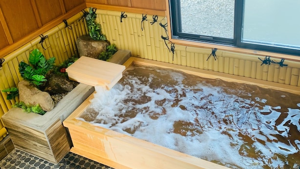 A hot tub with a wooden bench, surrounded by rocks and plants, with a window in the background. - Alloggio Nanki Shirahama (Shirahama)