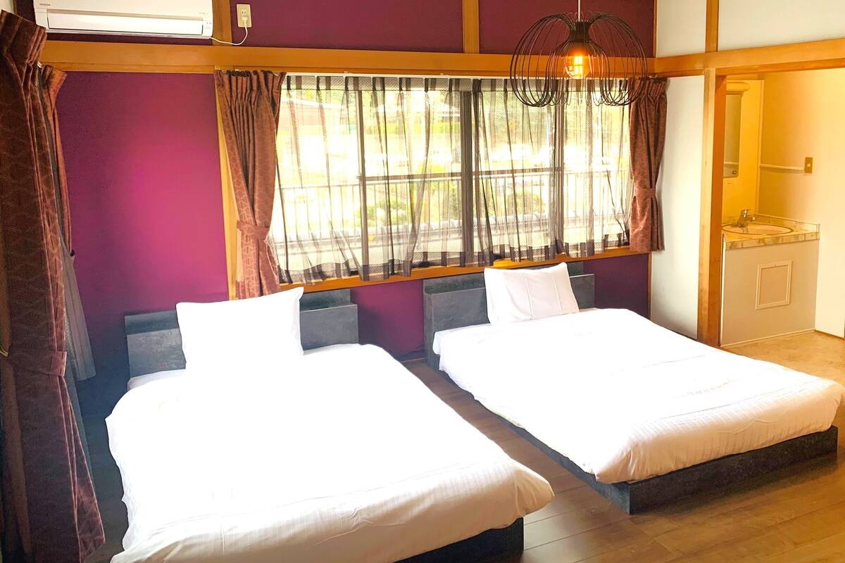 Een hotelkamer met twee bedden, een raam met gordijnen, een houten vloer, en een zichtbare badkamer door een open deur.