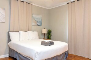 A neatly made bed with white linens, a lamp, and a plant on a bedside table in a room with beige curtains and a painting on the wall. - Pet-Friendly Home, Walk to Riverwalk, Free (Columbus)