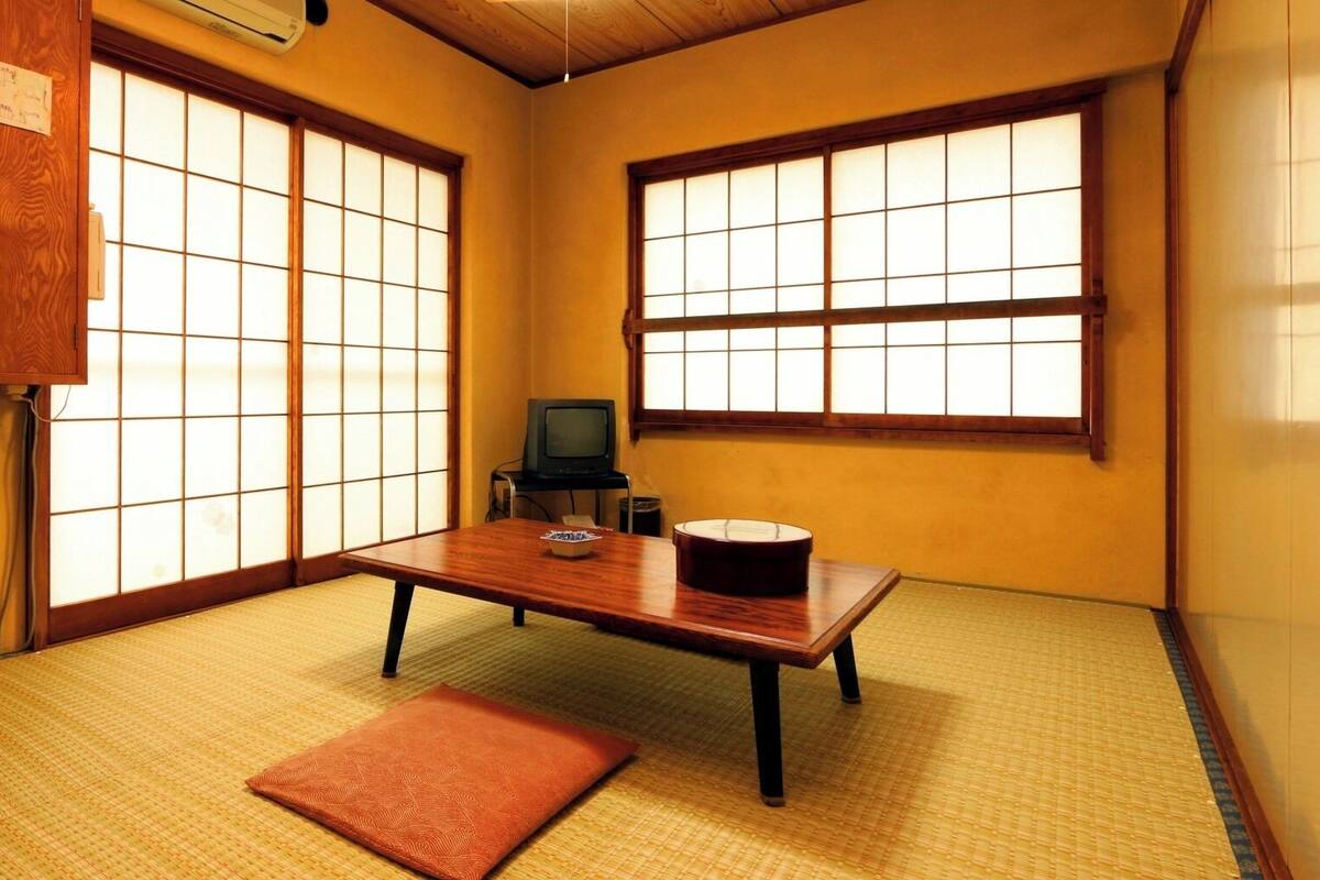 Ein traditioneller japanischer Raum mit Tatami-Boden, einem niedrigen Tisch, einem Fernseher und Schiebetüren.