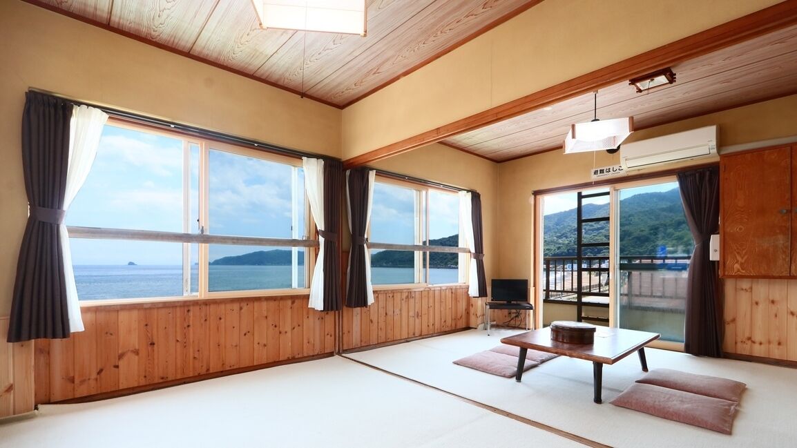 A room with wooden walls and floors, large windows with curtains, a tatami mat floor, a table with cushions, and a television.