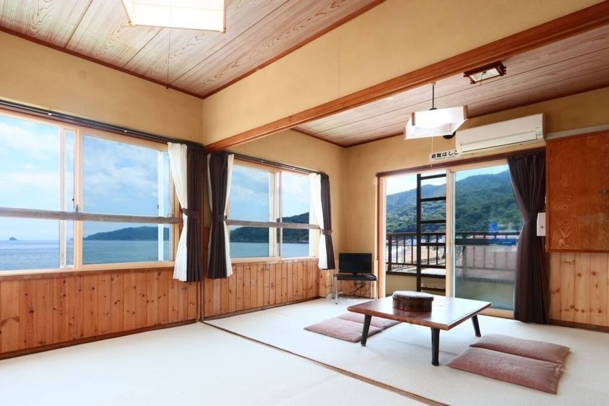 Ein Zimmer mit Holzwänden und -böden, großen fenstern mit Vorhängen, einem Tatami-Bodenbelag, einem Tisch mit Kissen und einem Fernseher.