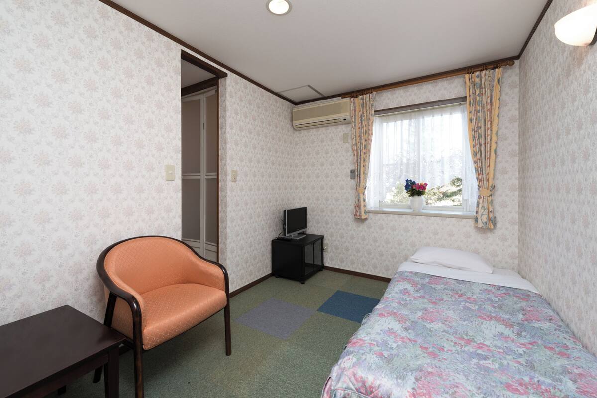 A room with a bed, a chair, a small table, a television on a stand, and a window with curtains.