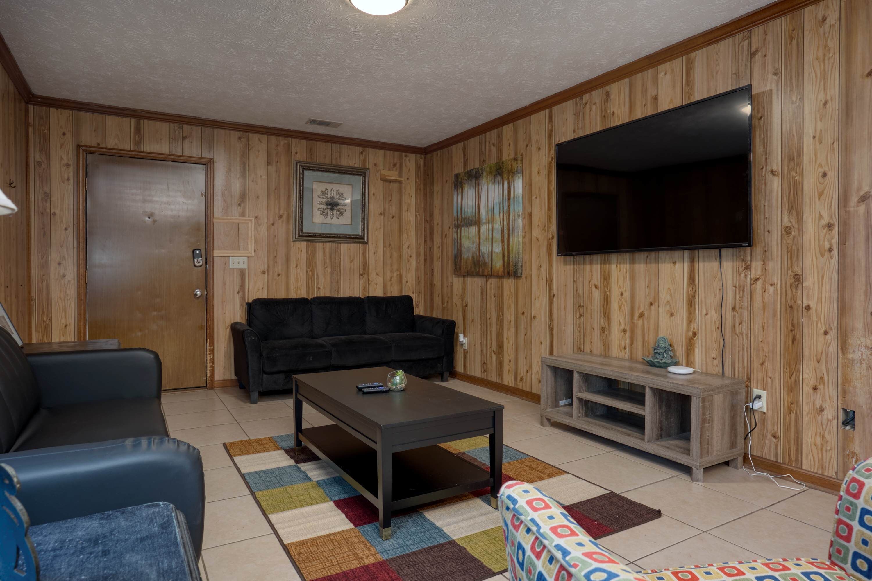 Un salon avec un mur lambrissé en bois, un canapé noir, une table basse et un téléviseur.