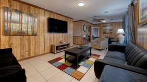 A living room with wooden paneling, a flat-screen TV, a coffee table, a sofa, a patterned rug, and a ceiling fan. - Mins to Fort Benning - The Columbus Cottage Guesthouse (Columbus)