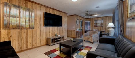Una sala de estar con paneles de madera, un televisor de pantalla plana, una mesa de centro, un sofá, una alfombra estampada y un ventilador de techo.
