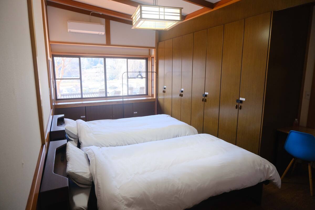 Quarto com duas camas, uma janela, um guarda-roupa e uma cadeira azul.