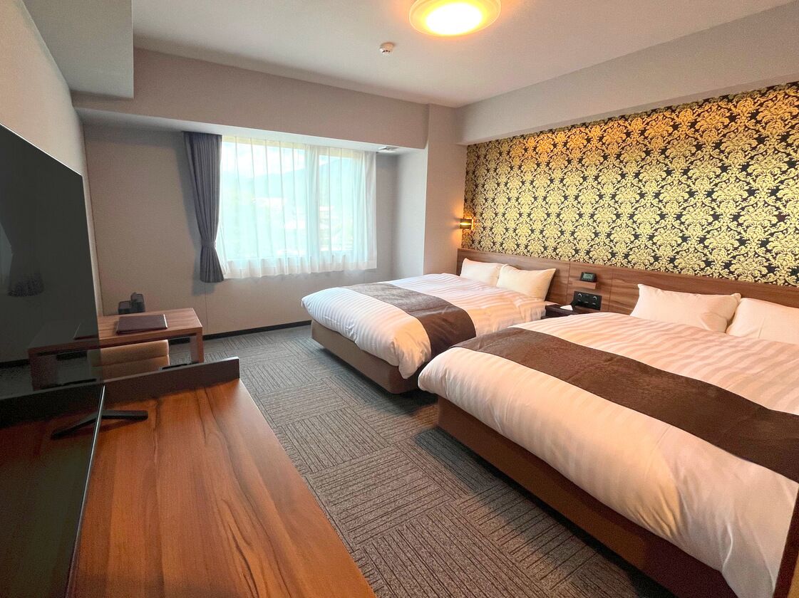 A hotel room with two beds, a desk, a TV, and a patterned wall.