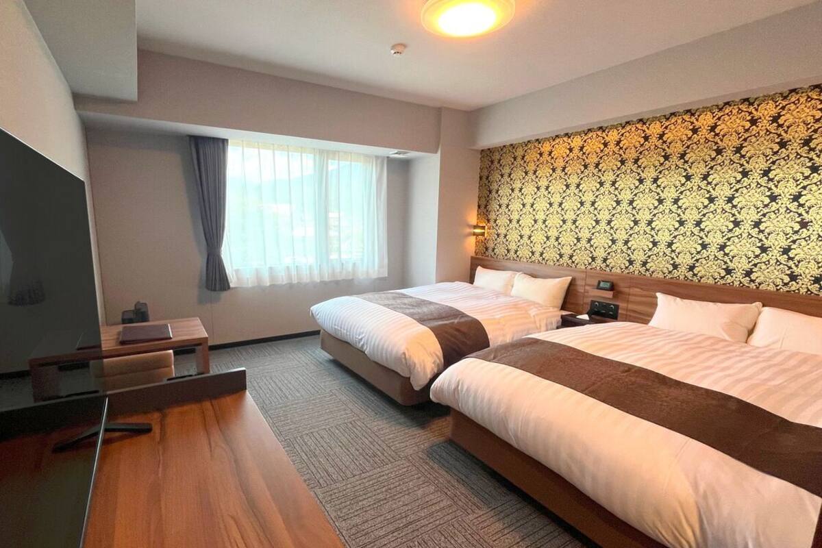 A hotel room with two beds, a desk, a TV, and a patterned wall.