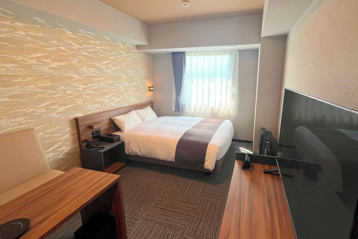 A hotel room with a bed, bedside table, TV, and a large window with curtains.