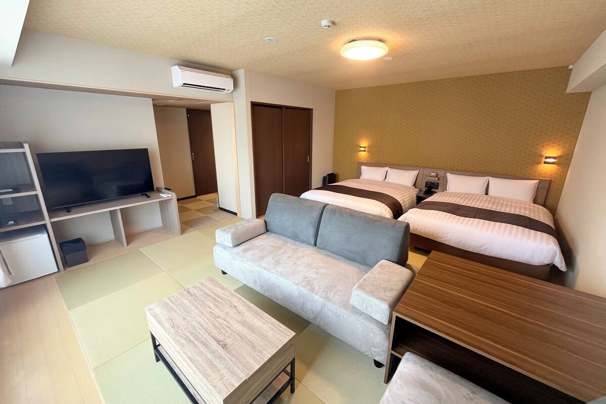 A modern hotel room with a flat-screen TV, a sofa, a wooden coffee table, and two beds with headboards.