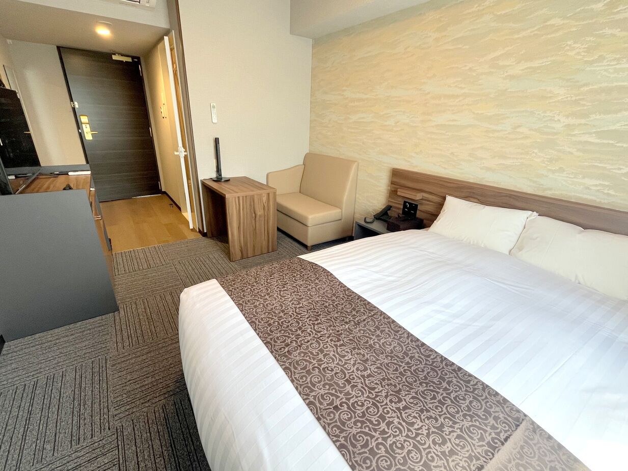A hotel room with a large bed, a desk, a chair, and a door leading to another room.