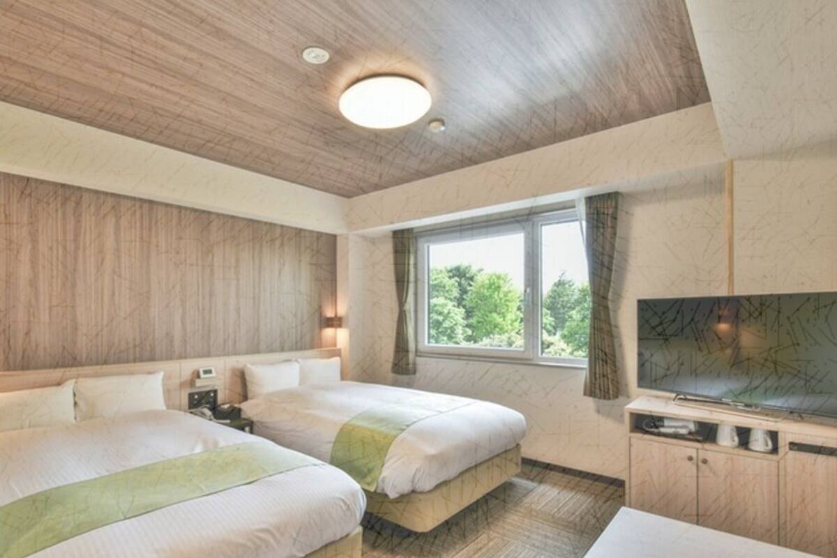 A hotel room with two beds, a TV, a window with curtains, and a wooden ceiling.