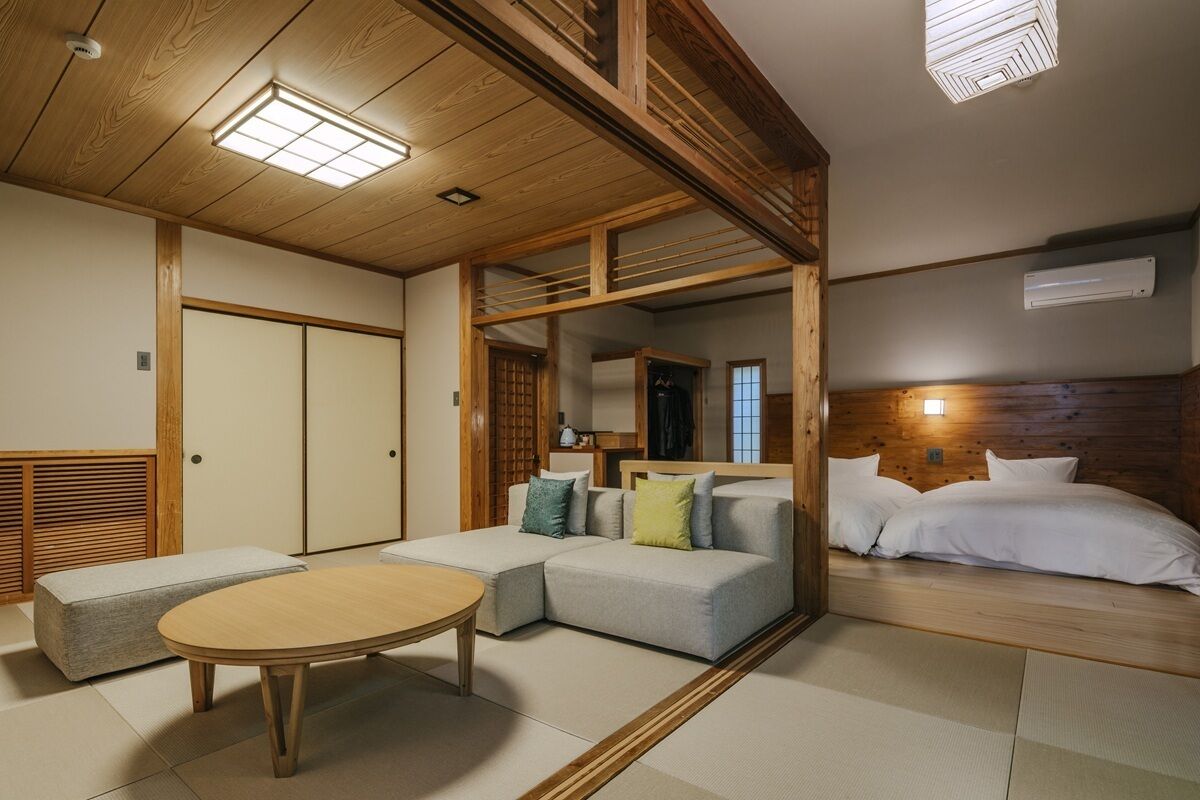 A traditional Japanese-style room with tatami flooring, a low wooden ceiling, a sofa, a bed, and a small round table.