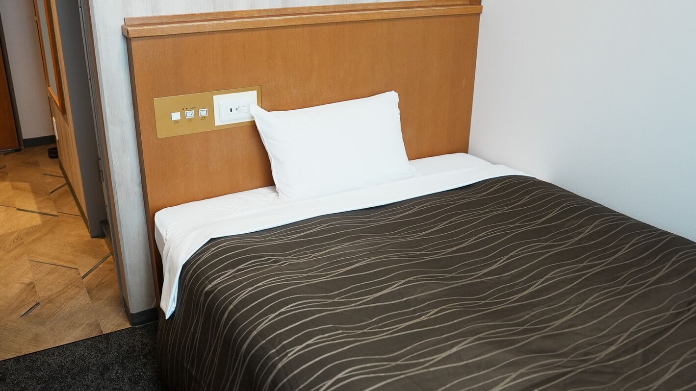 A hotel room with a wooden headboard, a white pillow, a brown patterned bedspread, and a power outlet on the wall.