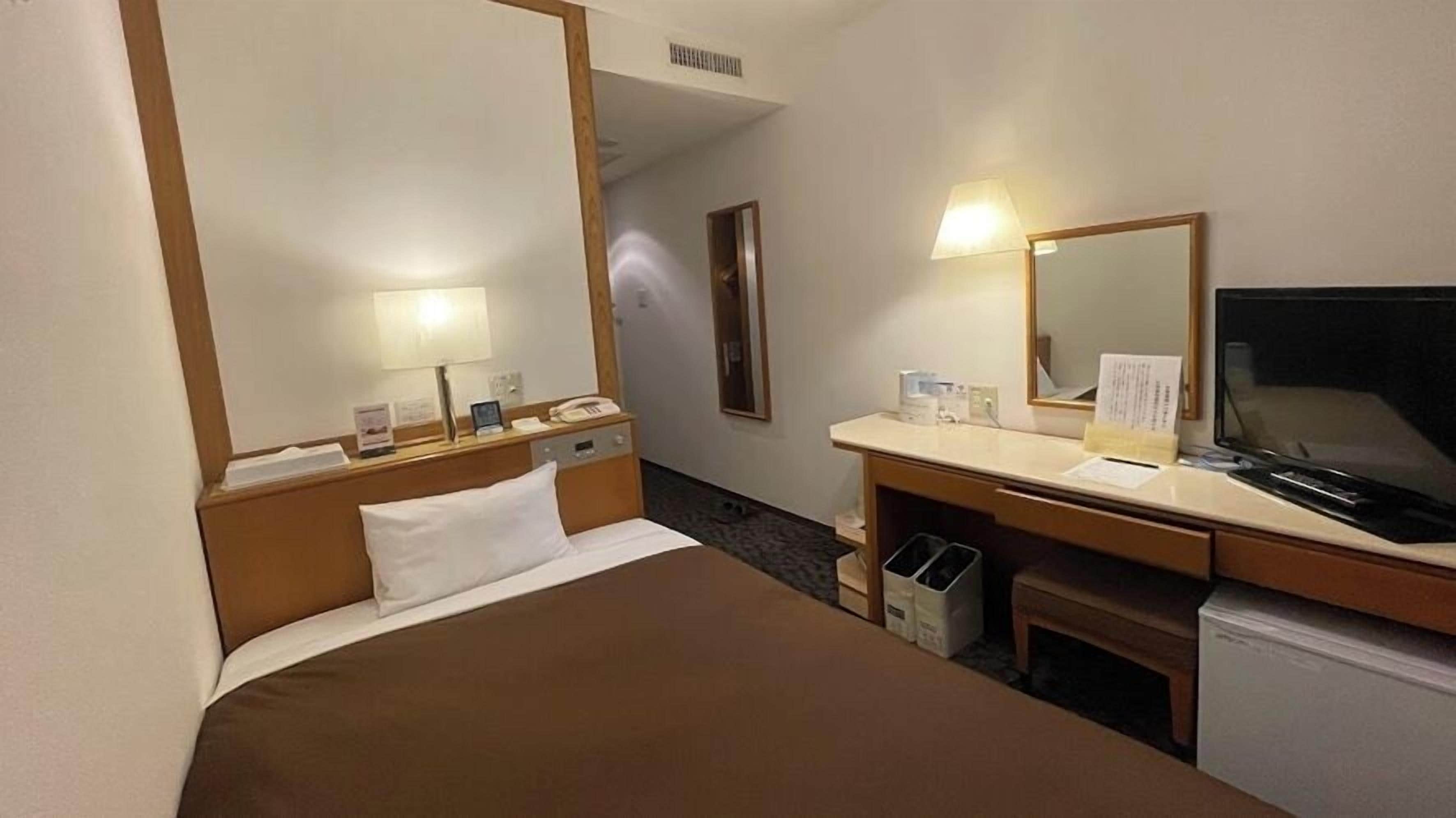 A hotel room with a bed, a desk with a lamp, a television, and a mirror.