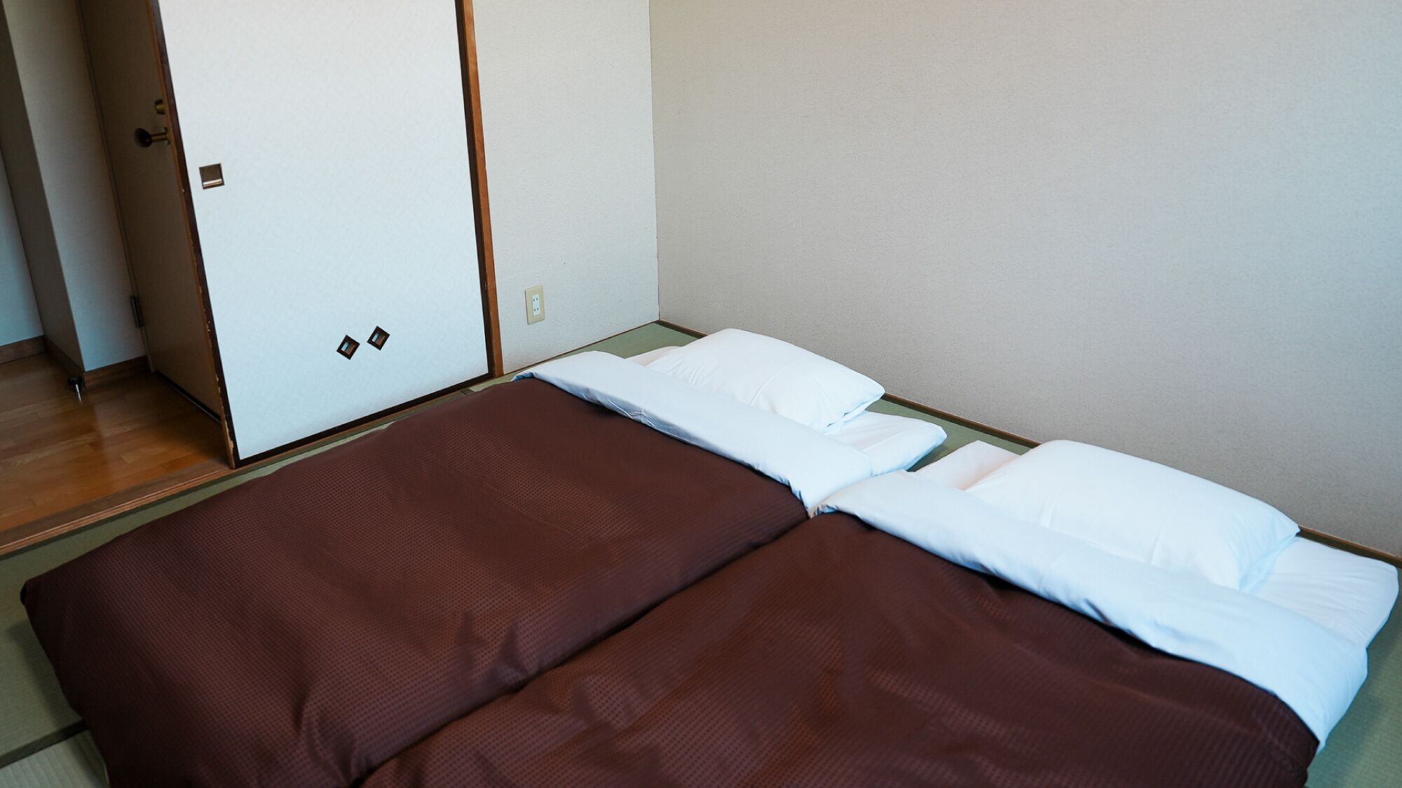 A single bed with a brown mattress and white pillows, a wooden floor, and a white wardrobe with a door.