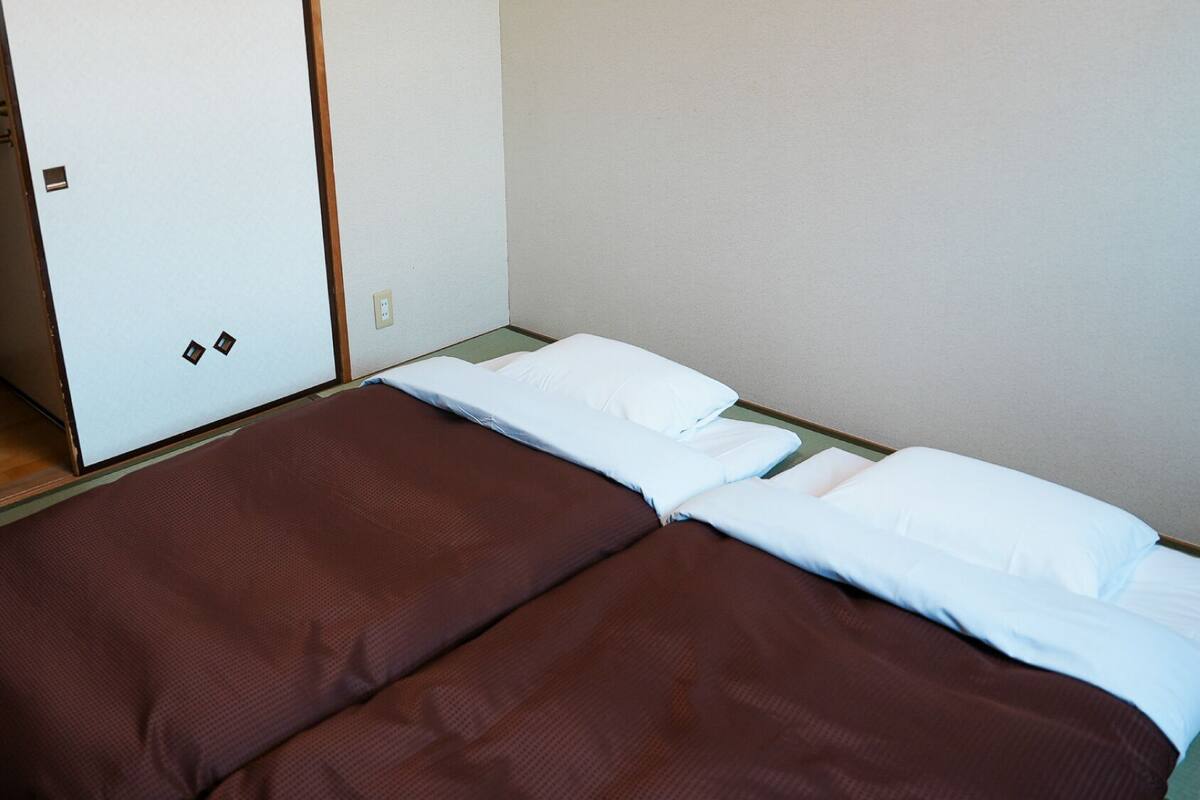 Una cama individual con colchón marrón y almohadas blancas, suelo de madera y un armario blanco con puerta.
