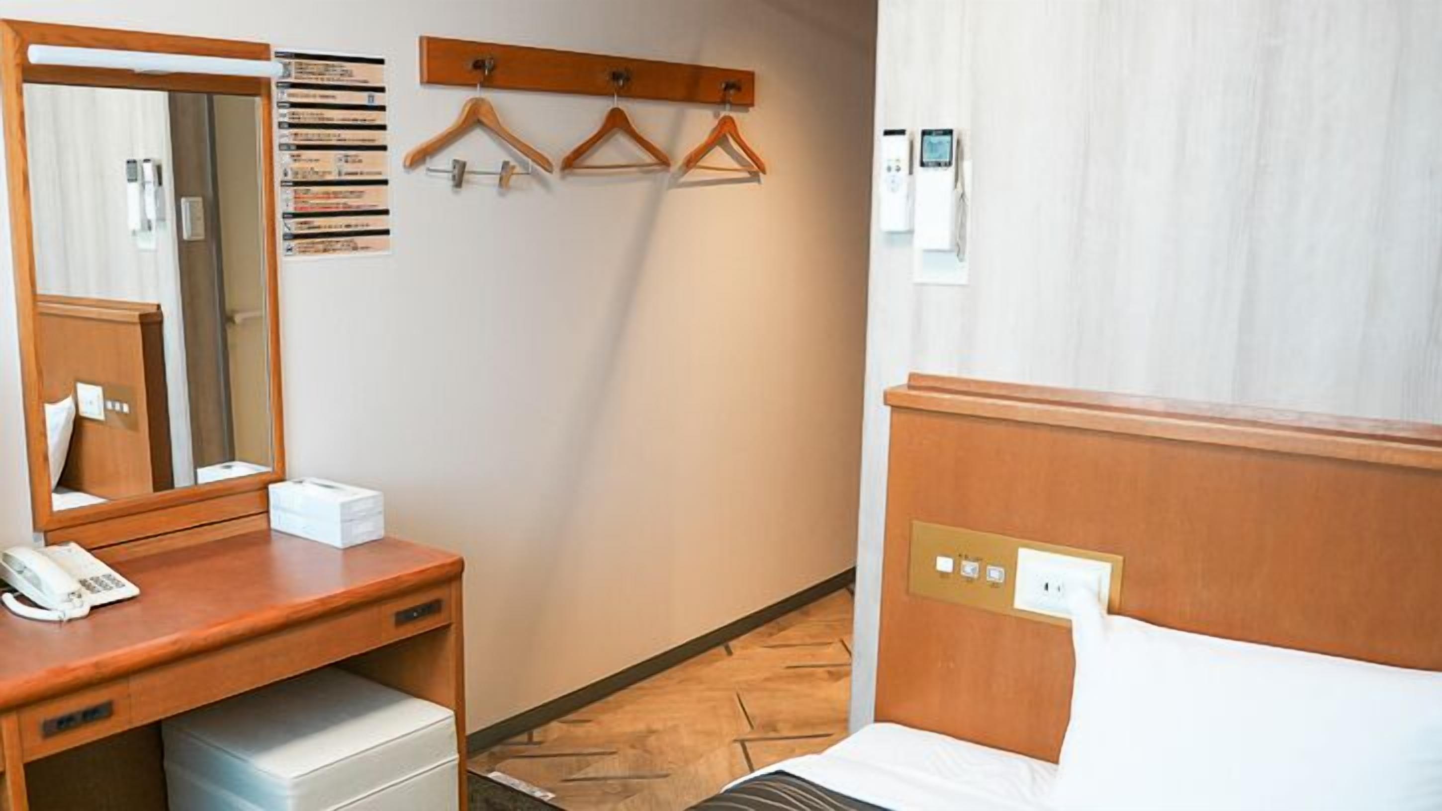 A hotel room with a wooden desk, a mirror, a telephone, a bedside table, a bed, a wall-mounted heater, and a clothing rack.