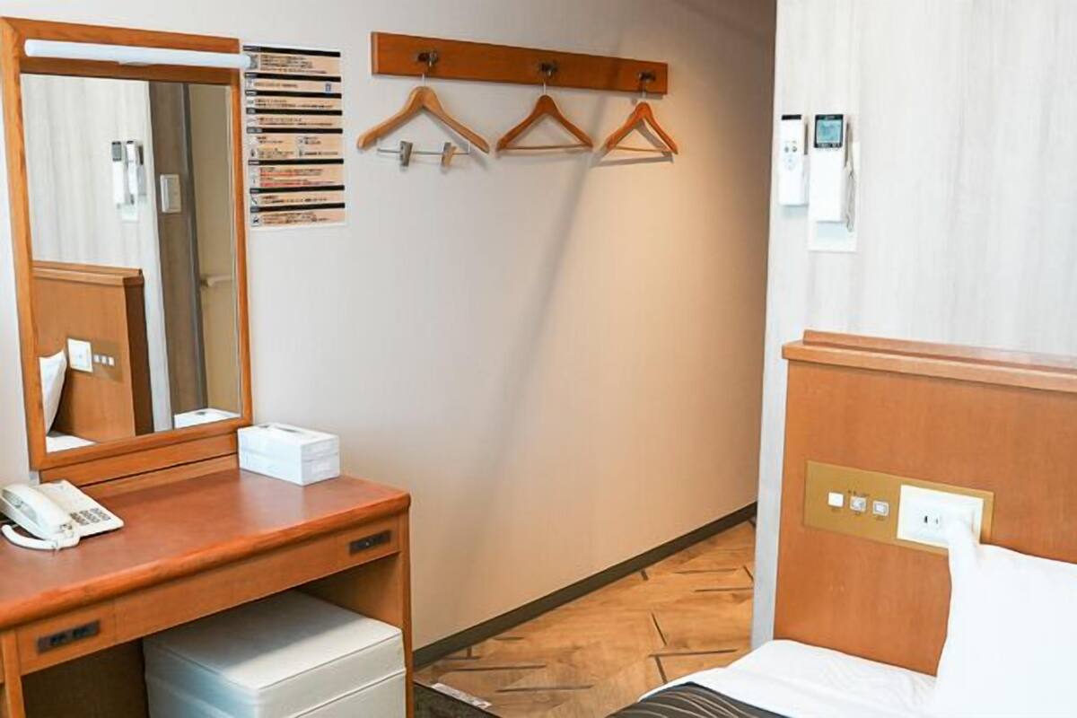 Habitación de hotel con escritorio de madera, espejo, teléfono, mesita de noche, cama, calentador de pared y perchero.