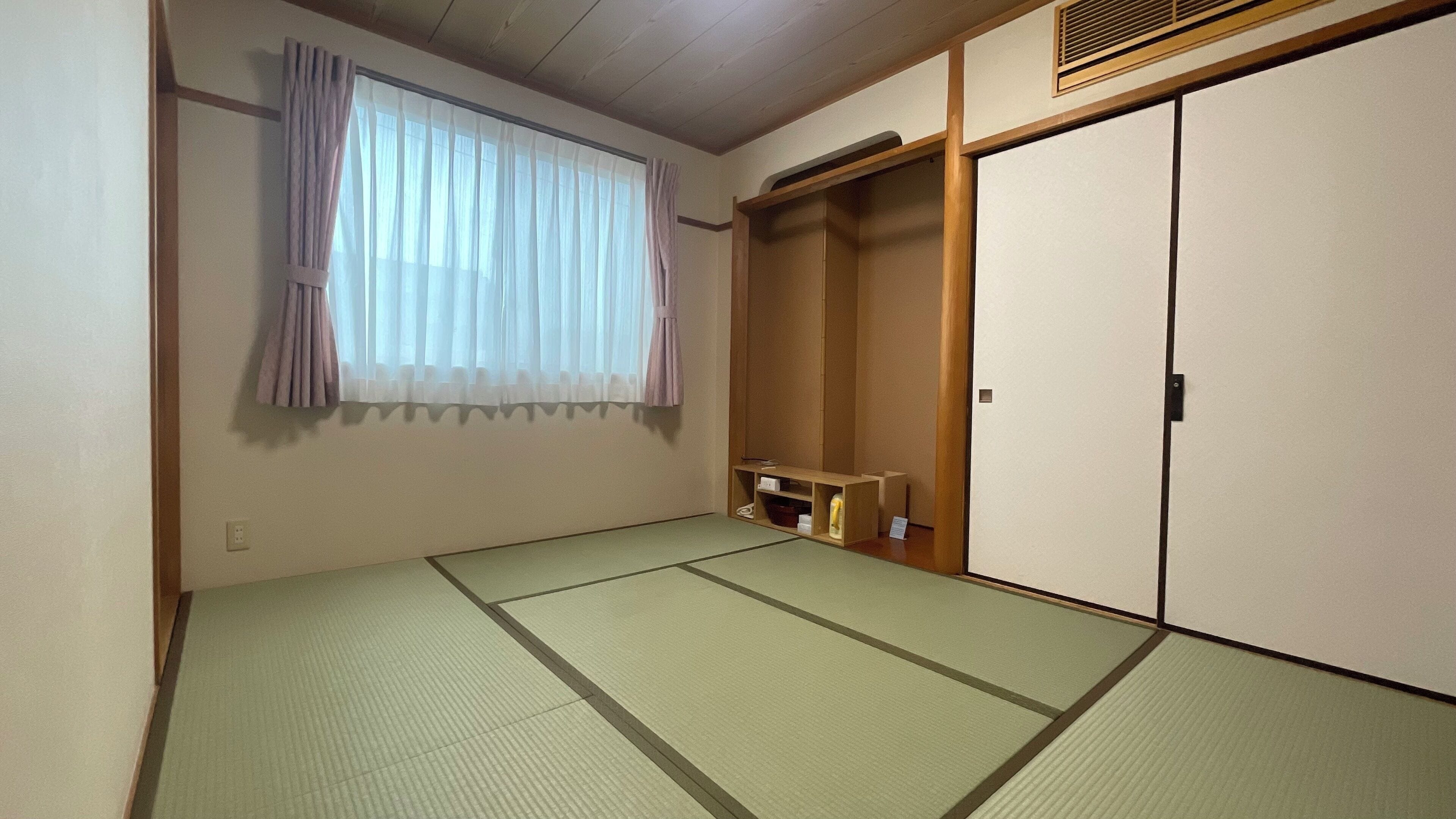A room with tatami flooring, a sliding door, a window with curtains, and a shelf with items.